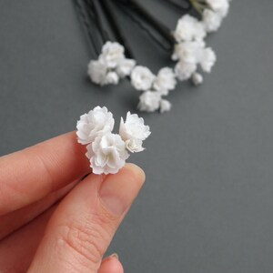May include: Three white flower hair pins, held in a hand, with a cluster of white flowers on each pin.