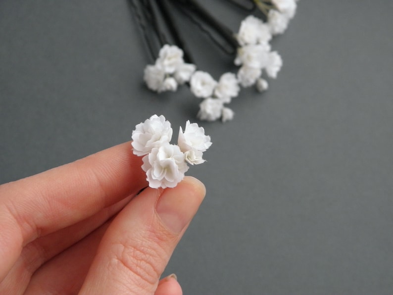 May include: Three white flower hair pins, held in a hand, with a cluster of white flowers on each pin.