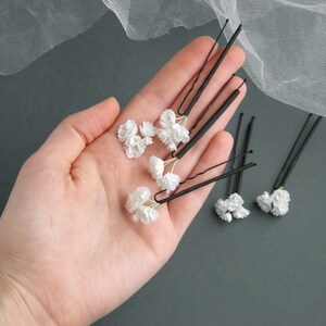 May include: Six black hair clips with white flower accents. The clips are arranged in a hand.