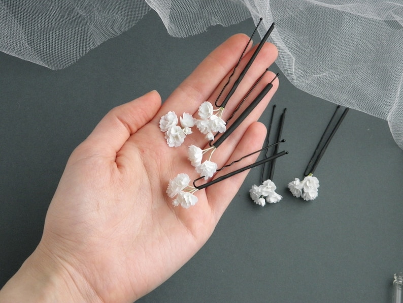 May include: Six black hair clips with white flower accents. The clips are arranged in a hand.