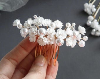 May include: A gold hair comb with white fabric flowers and pearl accents. The comb is held in a hand.