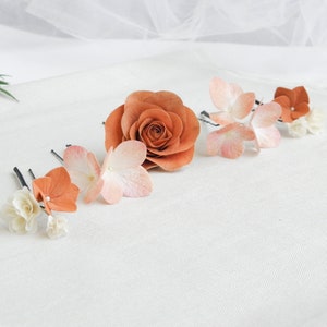 May include: A collection of floral hair accessories, including a large orange rose and several hair pins with orange, pink, and white flower accents. The flowers are arranged on a white surface, creating a delicate and elegant aesthetic.