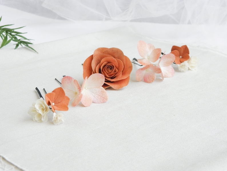 May include: A collection of floral hair accessories, including a large orange rose and several hair pins with orange, pink, and white flower accents. The flowers are arranged on a white surface, creating a delicate and elegant aesthetic.