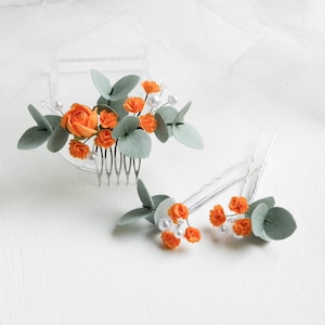 以下が含まれることがあります： オレンジと緑のフェルトの花と白いパールがあしらわれたヘアクリップ2個。大きなクリップにはコームが付いています。