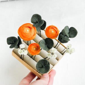 May include: A collection of orange ranunculus flower hair pins, dark green eucalyptus leaves, and small white flower hair pins arranged in a display box. The hair accessories are set against a neutral background.