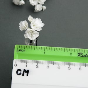 May include: A small bouquet of white paper flowers in a clear glass vase. The flowers are about 1 inch tall and the vase is about 1/2 inch tall. The flowers are arranged in a cluster and the vase is sitting on a gray surface.