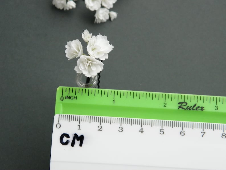 May include: A small bouquet of white paper flowers in a clear glass vase. The flowers are about 1 inch tall and the vase is about 1/2 inch tall. The flowers are arranged in a cluster and the vase is sitting on a gray surface.