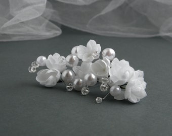 Puede incluir: Una delicada peineta floral blanca con flores de tela, perlas de imitación y detalles de cristal. El accesorio nupcial se encuentra sobre un fondo gris suave con un velo blanco transparente. Ideal para bodas y ocasiones especiales.