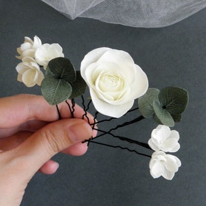 May include: Set of five white paper flower hair pins with green leaves. The flowers are arranged in a cluster and are attached to black bobby pins.