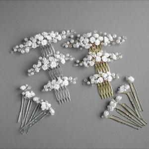 May include: A selection of bridal hair accessories, including combs and hairpins. The combs are adorned with white fabric flowers and faux pearls. The hairpins and combs are in silver and gold tones, set against a grey background.