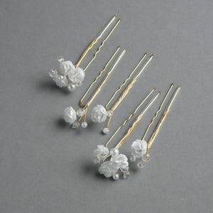 Può includere: Diverse forcine dorate con fiori di tessuto bianco, perle finte e dettagli in cristallo trasparente. Gli accessori per capelli sono disposti su una superficie grigia, ideali per matrimoni o eventi speciali.