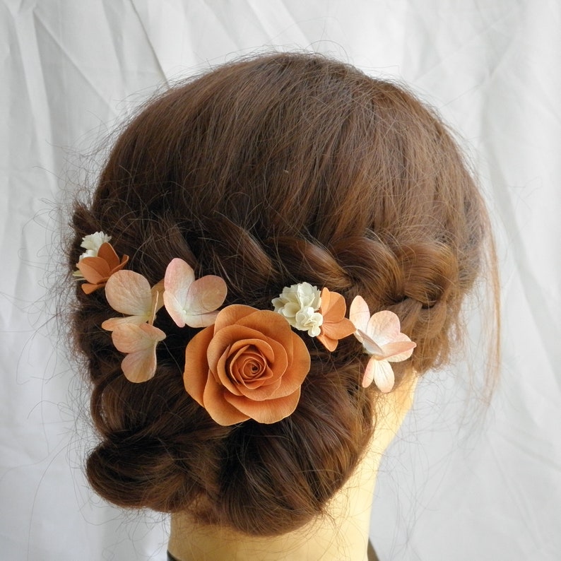 May include: A hair accessory featuring a large orange rose and smaller peach and white flowers. The floral arrangement is set against brown hair styled in a braid. The flowers are clustered together, creating a delicate and natural look.