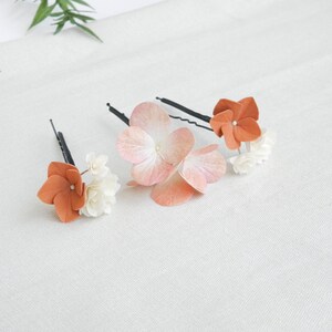 May include: Three floral hair pins. Each pin features a cluster of small white flowers and a larger, orange-colored flower. One pin has a large, light pink flower. The pins are on a white surface.