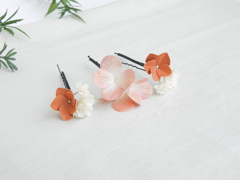 May include: Three floral hair pins. Each pin features a cluster of small white flowers and a larger, orange-colored flower. One pin has a large, light pink flower. The pins are on a white surface.