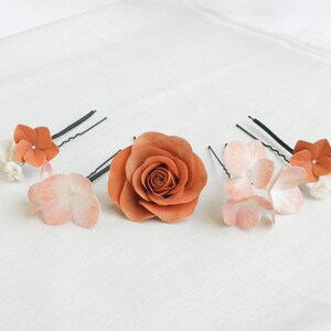 May include: A collection of floral hair accessories, including a large rust-colored rose and several hairpins adorned with smaller flowers in shades of peach, white, and rust. The items are arranged on a white fabric surface.