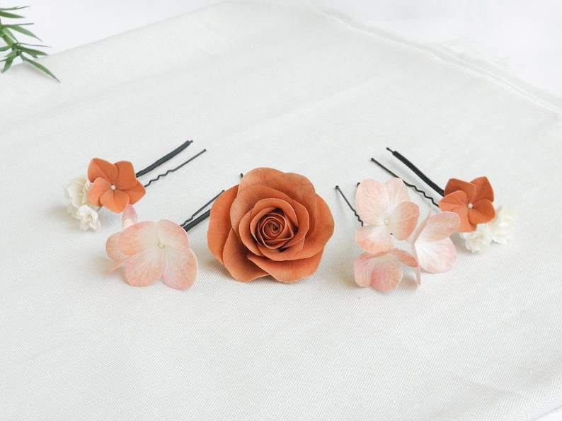 May include: A collection of floral hair accessories, including a large rust-colored rose and several hairpins adorned with smaller flowers in shades of peach, white, and rust. The items are arranged on a white fabric surface.