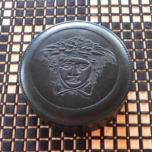 May include: A circular black leather case featuring a debossed Medusa head emblem on the lid. The lid is secured with a stitched border around the perimeter, showcasing a detailed relief of the mythological figure with flowing hair.