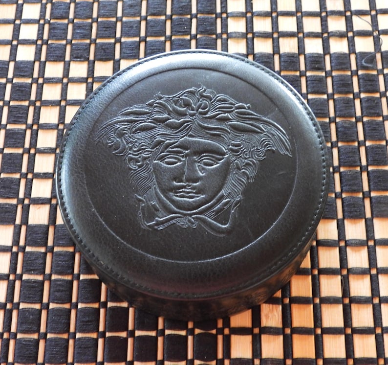 May include: A circular black leather case featuring a debossed Medusa head emblem on the lid. The lid is secured with a stitched border around the perimeter, showcasing a detailed relief of the mythological figure with flowing hair.