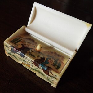 May include: A rectangular off-white box with a hinged lid that is open. The front panel features a hand-painted scene of two figures on horseback, one holding a bow and arrow, rendered in muted earth tones, blues, and greens with a small knob.