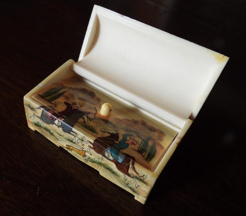 May include: A rectangular off-white box with a hinged lid that is open. The front panel features a hand-painted scene of two figures on horseback, one holding a bow and arrow, rendered in muted earth tones, blues, and greens with a small knob.