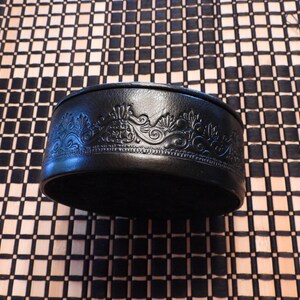 May include: A round black container with a smooth, dark surface featuring an intricate, embossed decorative pattern around the side. The design consists of repeating floral and scroll-like motifs that are pressed into the material, creating a textured relief effect along the entire circumference of the circular object.