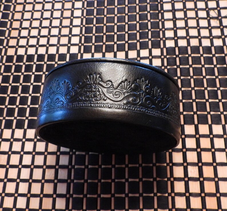 May include: A round black container with a smooth, dark surface featuring an intricate, embossed decorative pattern around the side. The design consists of repeating floral and scroll-like motifs that are pressed into the material, creating a textured relief effect along the entire circumference of the circular object.