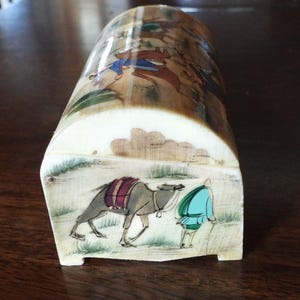 May include: A curved, off-white box featuring hand-painted scenes. The front panel displays a person in a teal tunic walking beside a grey camel with a purple saddle. The top surface shows a detailed landscape with figures in blue and brown clothing.