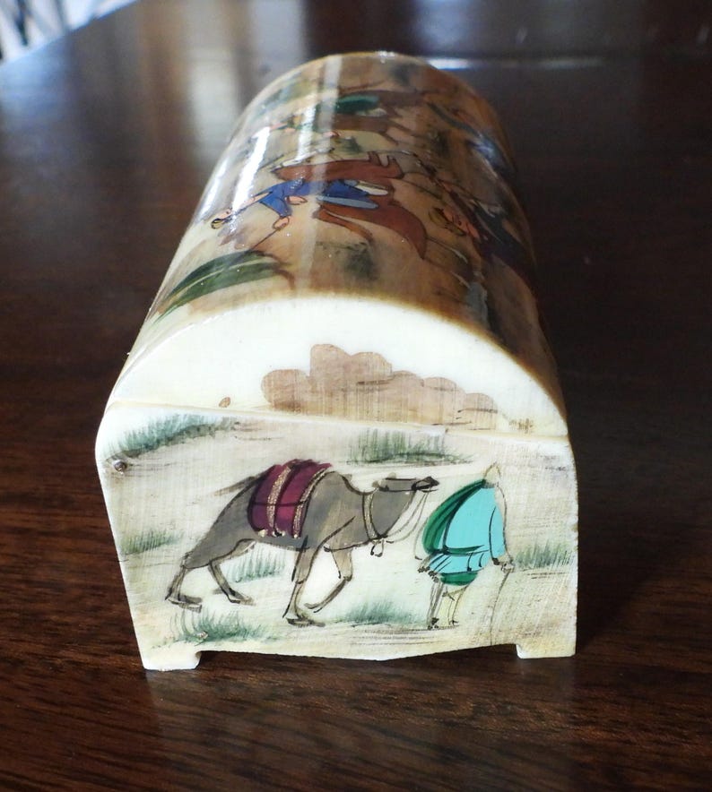 May include: A curved, off-white box featuring hand-painted scenes. The front panel displays a person in a teal tunic walking beside a grey camel with a purple saddle. The top surface shows a detailed landscape with figures in blue and brown clothing.