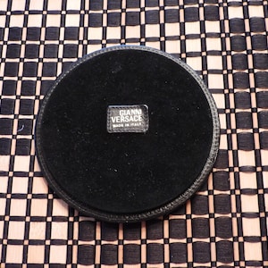 May include: A circular black coaster with a smooth leather-like border and a soft, dark fabric centre. A small rectangular black label is stitched in the middle, featuring the white text GIANNI VERSACE and the words MADE IN ITALY below it.
