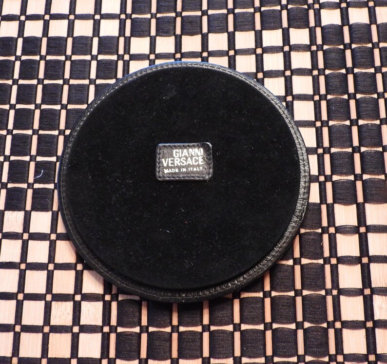 May include: A circular black coaster with a smooth leather-like border and a soft, dark fabric centre. A small rectangular black label is stitched in the middle, featuring the white text GIANNI VERSACE and the words MADE IN ITALY below it.
