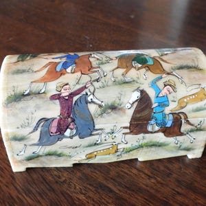 May include: A curved, rectangular box with an off-white base color, featuring a detailed hand-painted scene of four figures on horseback engaged in a hunt. The riders wear colorful tunics and hats, while galloping across a landscape with small animals.