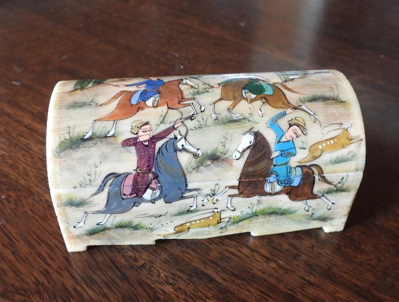 May include: A curved, rectangular box with an off-white base color, featuring a detailed hand-painted scene of four figures on horseback engaged in a hunt. The riders wear colorful tunics and hats, while galloping across a landscape with small animals.