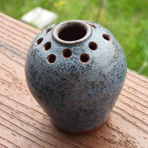 May include: A round ceramic flower frog vase with a speckled blue and gray glaze. The vessel features a central opening surrounded by a ring of smaller circular holes on the top surface, designed for arranging individual flower stems.