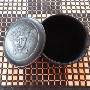 May include: A round black leather jewellery box with a removable lid. The lid features an embossed Medusa head design in the centre. The interior of the box is lined with black velvet, providing a soft surface for storing small items or accessories.