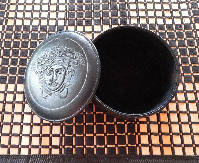 May include: A round black leather jewellery box with a removable lid. The lid features an embossed Medusa head design in the centre. The interior of the box is lined with black velvet, providing a soft surface for storing small items or accessories.