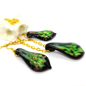 May include: A set of three glass pendants with gold chains. Each pendant is teardrop-shaped, with a black base and green floral design. Gold flecks and accents are visible within the glass. The pendants are displayed on a white surface.