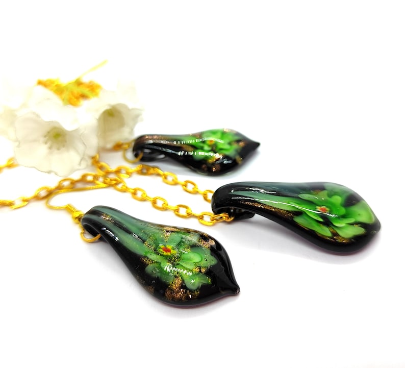 May include: A set of three glass pendants with gold chains. Each pendant is teardrop-shaped, with a black base and green floral design. Gold flecks and accents are visible within the glass. The pendants are displayed on a white surface.
