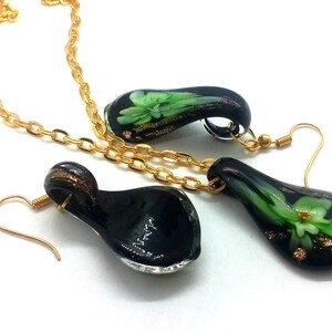 May include: A jewelry set featuring a necklace and earrings. The set includes a gold chain necklace and matching earrings with a teardrop-shaped pendant. The pendants are black with green and gold floral designs.