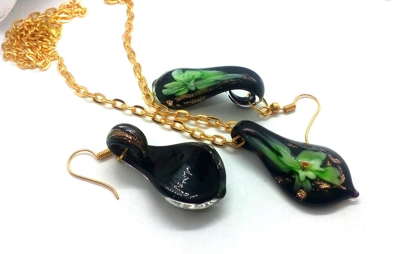 May include: A jewelry set featuring a necklace and earrings. The set includes a gold chain necklace and matching earrings with a teardrop-shaped pendant. The pendants are black with green and gold floral designs.