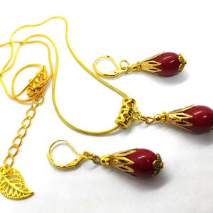 Pu&ograve; includere: Un set di gioielli color oro composto da una collana e due orecchini abbinati. Ogni pezzo presenta una perla rossa liscia a forma di goccia racchiusa in un cappuccio di metallo dorato decorato. La collana include una catenina sottile con un ciondolo a forma di foglia.