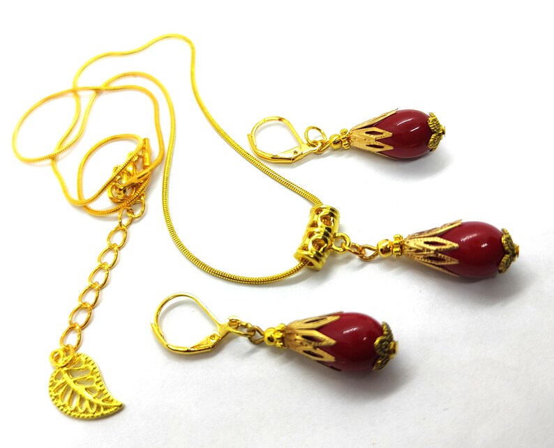 Pu&ograve; includere: Un set di gioielli color oro composto da una collana e due orecchini abbinati. Ogni pezzo presenta una perla rossa liscia a forma di goccia racchiusa in un cappuccio di metallo dorato decorato. La collana include una catenina sottile con un ciondolo a forma di foglia.