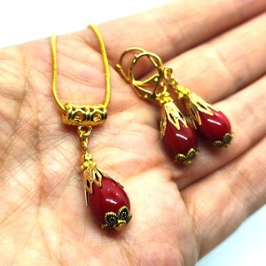 Pu&ograve; includere: Una collana e un paio di orecchini pendenti abbinati in tonalit&agrave; oro. Ogni pezzo presenta una pietra rossa liscia a forma di goccia, racchiusa in un cappuccio di metallo dorato decorato con una base ispirata a motivi floreali.