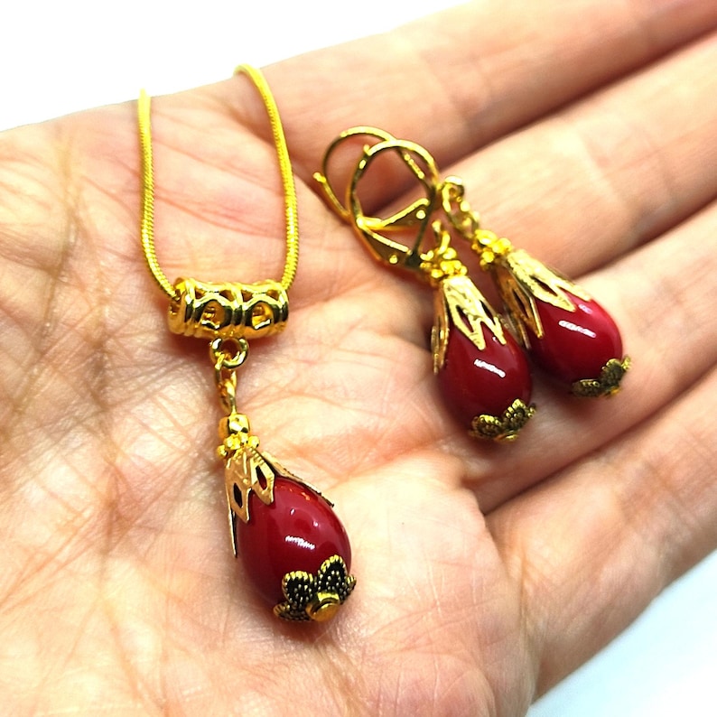 Pu&ograve; includere: Una collana e un paio di orecchini pendenti abbinati in tonalit&agrave; oro. Ogni pezzo presenta una pietra rossa liscia a forma di goccia, racchiusa in un cappuccio di metallo dorato decorato con una base ispirata a motivi floreali.