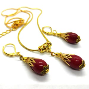 Pu&ograve; includere: Un set di gioielli color oro composto da una collana e due orecchini abbinati. Ogni pezzo presenta una perla rossa liscia a forma di goccia, racchiusa in un cappuccio decorativo in metallo dorato. La collana include un distanziatore cilindrico.