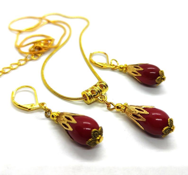 Pu&ograve; includere: Un set di gioielli color oro composto da una collana e due orecchini abbinati. Ogni pezzo presenta una perla rossa liscia a forma di goccia, racchiusa in un cappuccio decorativo in metallo dorato. La collana include un distanziatore cilindrico.