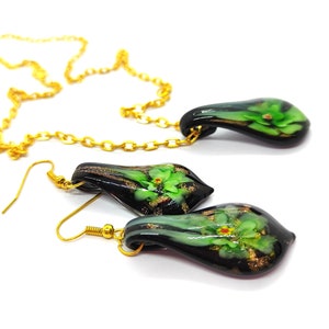 May include: A gold chain necklace and matching earrings set. The pendant and earrings are teardrop-shaped, with a black base and green floral design. Gold accents and hooks complete the jewelry set.