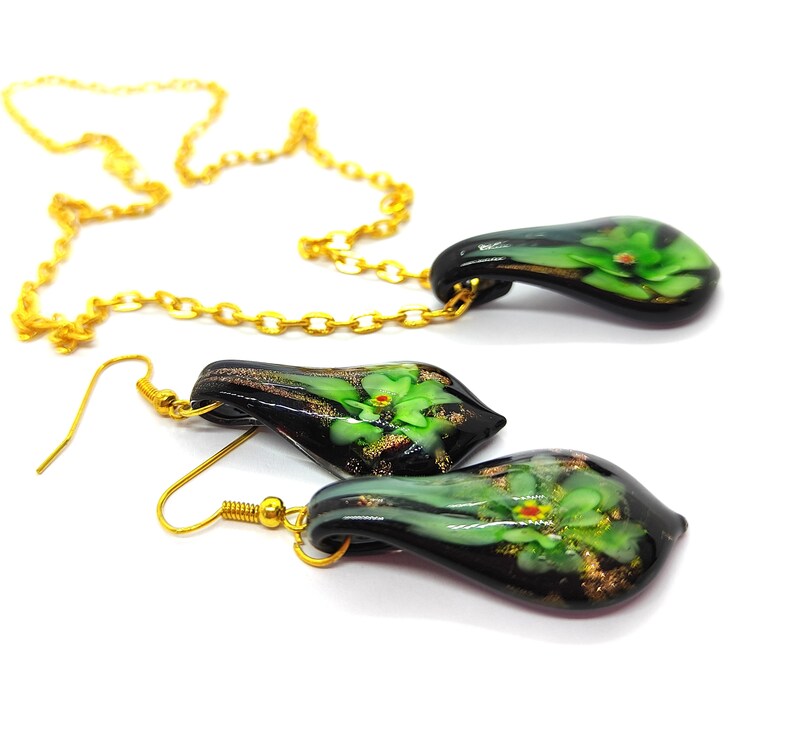May include: A gold chain necklace and matching earrings set. The pendant and earrings are teardrop-shaped, with a black base and green floral design. Gold accents and hooks complete the jewelry set.