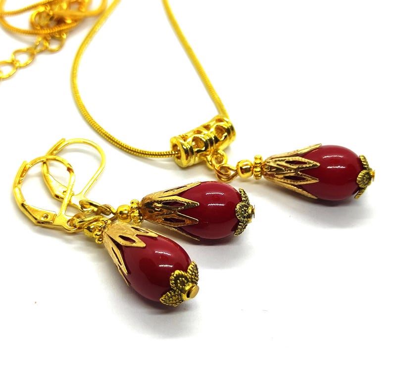 Pu&ograve; includere: Una collana e un paio di orecchini pendenti abbinati in metallo dorato, ciascuno caratterizzato da una perla liscia rosso scuro a forma di goccia, racchiusa in una capsula di metallo dorato lavorata a filigrana. La collana ha una catena sottile.