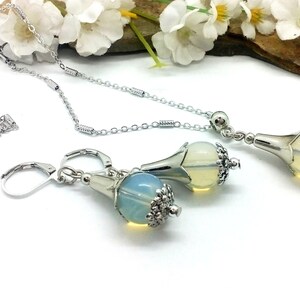 Puede incluir: Un conjunto de collar y pendientes de plata con un colgante en forma de l&aacute;grima y pendientes a juego. El colgante y los pendientes presentan una piedra clara e iridiscente similar al &oacute;palo engastada en un engaste de plata en forma de flor.