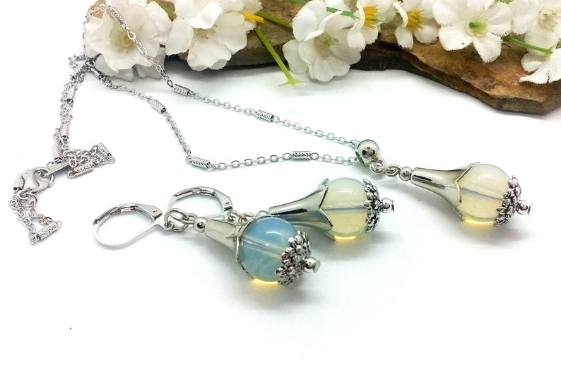 Puede incluir: Un conjunto de collar y pendientes de plata con un colgante en forma de l&aacute;grima y pendientes a juego. El colgante y los pendientes presentan una piedra clara e iridiscente similar al &oacute;palo engastada en un engaste de plata en forma de flor.
